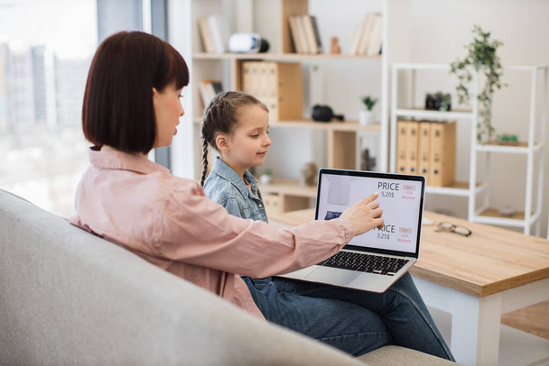 Caring mother with little daughter browsing ecommerce site for searching discount of new trendy apparel. Young woman and pretty girl sitting together on comfy couch and using modern wireless laptop. - Foto, immagini
