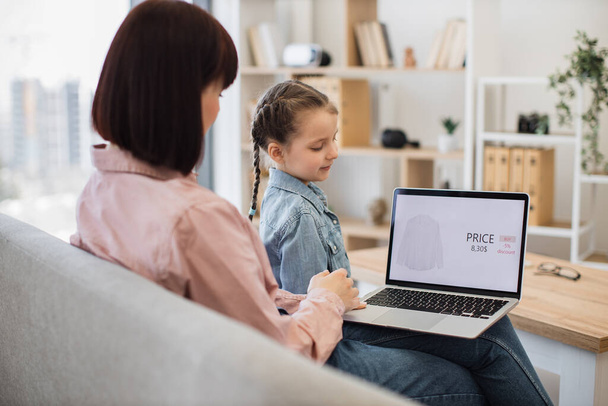 Dark haired caucasian woman and pretty little girl choosing clothes together online during sale season. Mother with daughter using portable laptop for shopping while enjoying time at home. - Foto, Bild