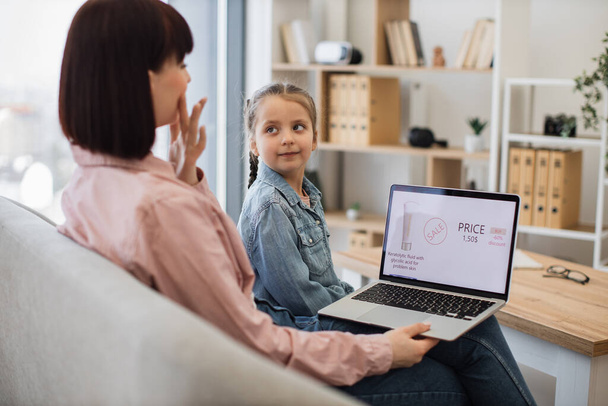 Rear view of beautiful woman holding laptop and cute girl looking over shoulder at her mom. Young females discussing facial beauty products provided at big discounts in online cosmetics webstore. - Foto, Bild