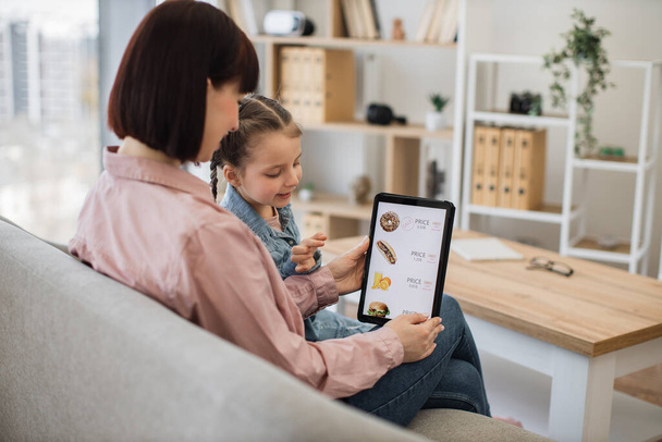 Relaxed mother and daughter searching e-commerce offers on restaurant food with tablet application while having fun indoors. Efficient online clients seizing moment of reduced prices on products. - Foto, Imagen