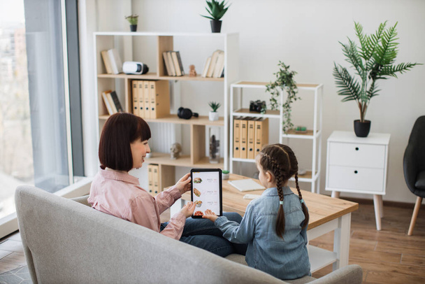 Happy mother and daughter resting on couch and pointing with their hands at tablet screen where junk food service online app being shown. Cheerful female shoppers making use of e-commerce. - Foto, Bild