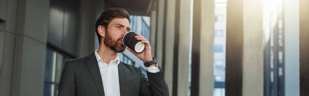 Attractive businessman in suit drinking coffee near office building during break. Blurred background - Fotografie, Obrázek