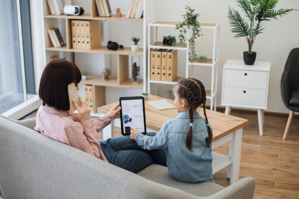 Brunette caucasian woman talking on phone and holding modern gadget in hand while little girl sitting next to her. Young ladies ordering new casual outfit at sales on virtual clothes store website. - Fotó, kép