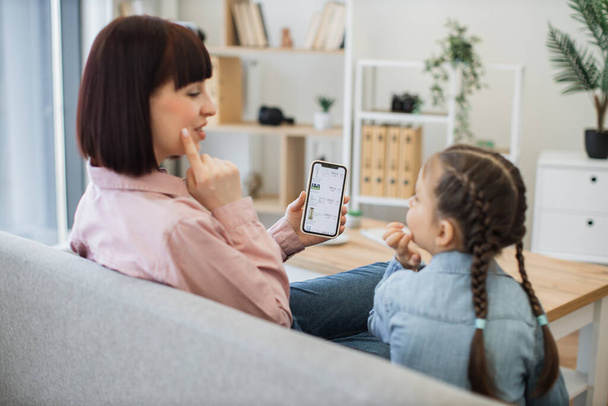 Beautiful young mother teaching her little daughter main rules of choosing cosmetics while browsing online boutique during sale season. Happy family having fun while doing home shopping on smartphone. - Foto, Bild