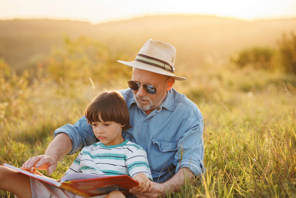 Little boy and his grandfather sitting on a grass and reading a book in the field. Boy and grandfather spending time together at summer. Brunette boy wearing striped t-shirt and man blue shirt and - Foto, Bild