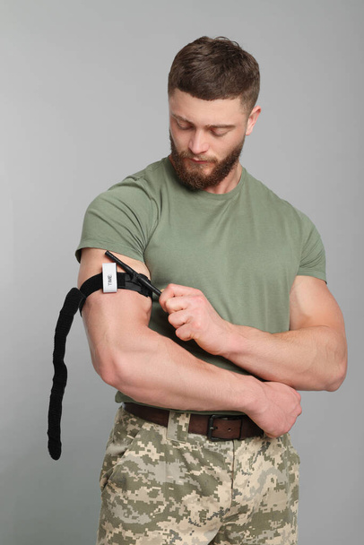 Soldier in military uniform applying medical tourniquet on arm against light grey background - Foto, imagen