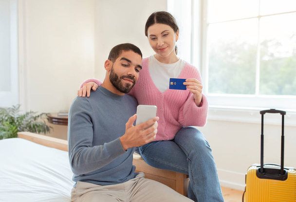 Tourists Couple Using Smartphone And Credit Card Booking Hotel Room Sitting With Travel Suitcase In Modern Bedroom At Home. Spouses Paying For Accomodation Via Mobile Application Concept - Photo, image