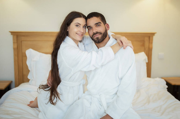 Happy Diverse Spouses Embracing Sitting On Bed Indoors. Shot Of Loving Couple Posing In Modern Bedroom At Home, Wearing White Bathrobes. Marriage And Romantic Relationship Concept - Foto, afbeelding
