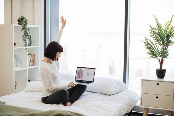Caucasian woman with brown hair sitting on bed cheerfully holding hands up while looking on laptop screen. Female customer feeling overjoyed while buying apparel with big discount. - Foto, Bild