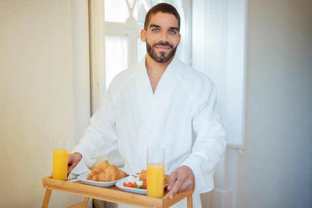 Desayuno romántico en la cama. Guapo hombre de Oriente Medio sosteniendo bandeja de mesa con cruasanes, ensalada y jugo de naranja sonriendo a la cámara de pie en la habitación de hotel interior, con albornoz blanco - Foto, imagen