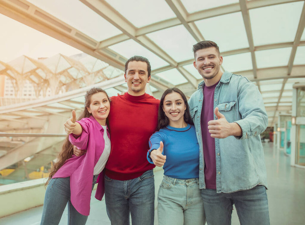 We Like Traveling. Four Friends Gesturing Thumbs Up Approving Vacation Offer Smiling To Camera Standing In Airport Indoors. Two Travelers Couples Showing Like Together. Transportation Concept - Photo, Image