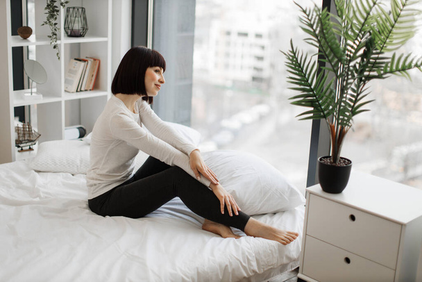 Attractive caucasian woman in domestic outfit resting on white bed with panoramic windows on background. Leisure time at home and lifestyles concept. - Photo, image