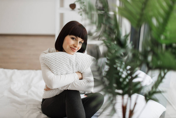 Adorable dark haired woman sitting on comfy bed with embrace of soft pillow, smiling and looking at camera. Caucasian female in casual attire enjoying leisure time alone at cozy home. - Фото, изображение