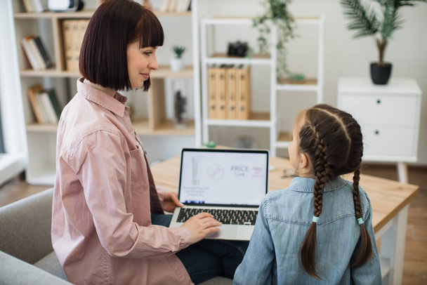 Close up of caucasian woman and little girl sitting on couch with modern laptop and searching available discounts on makeup products. Online store with cosmetics on sale on computer screen. - Foto, Imagem
