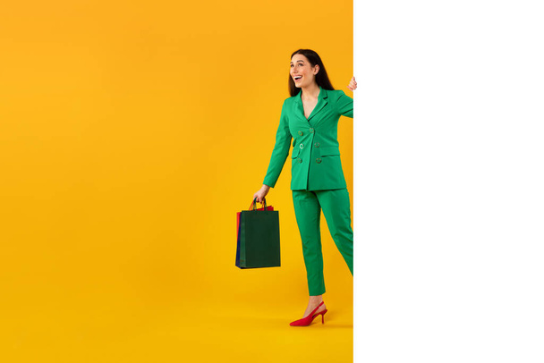 Glückliche stylische Dame mit Einkaufstaschen, die aus einem leeren weißen Papierbanner mit Platz für Werbung auf gelbem Studiohintergrund lugt, freier Platz. Mockup für Ihr Anzeigendesign - Foto, Bild