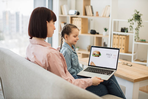Attractive dark haired woman sitting on couch with her lovely daughter and choosing fast food with discount on website. Caucasian family of two using modern laptop for ordering takeaway lunch. - Фото, изображение