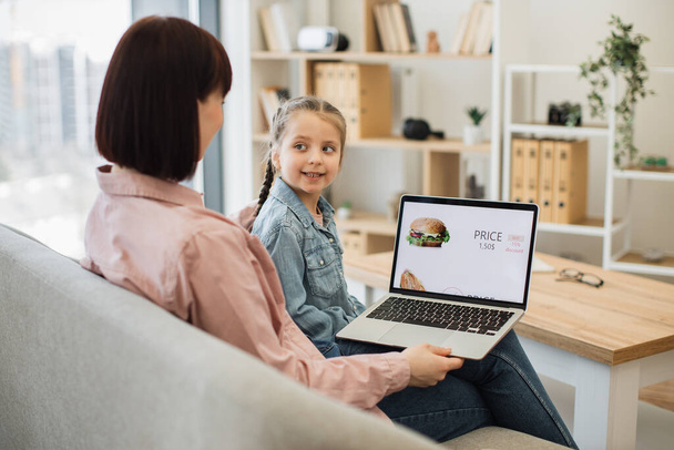 Attractive dark haired woman sitting on couch with her lovely daughter and choosing fast food with discount on website. Caucasian family of two using modern laptop for ordering takeaway lunch. - Foto, immagini