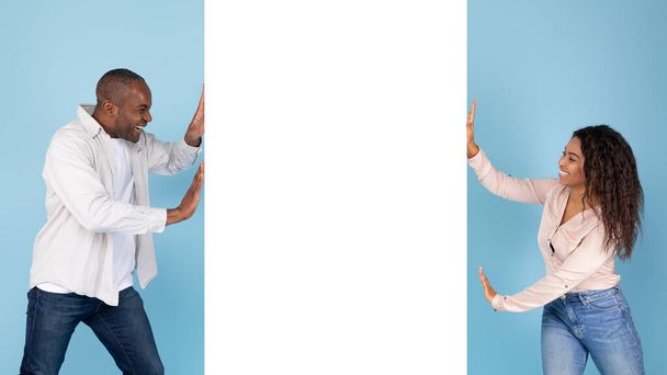 Возбужденная африканская пара, толкающая белый пустой вертикальный рекламный плакат, демонстрирующий место для вашего дизайна, синий студийный фон, баннер, панорама - Фото, изображение