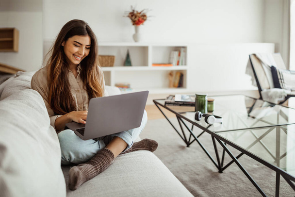 Smiling busy pretty young asian female typing on laptop in living room interior. Meeting remotely, work, business, freelance at home, new normal with technology and social networks - 写真・画像