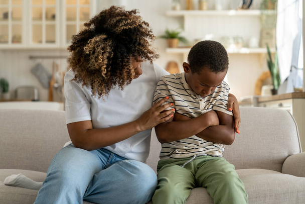 Loving african american mother embracing hugging sad little son supporting child, mom parent trying to make peace with offended upset kid, sitting together on sofa at home. Parent-child conflict - Foto, Bild
