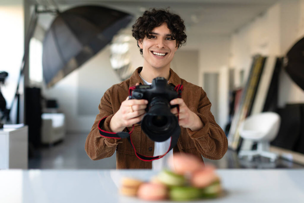 Νεαρό αρσενικό φωτογράφος τροφίμων χρησιμοποιώντας ψηφιακή φωτογραφική μηχανή και τη λήψη φωτογραφιών του επιδορπίου, κοιτάζοντας και χαμογελώντας στην κάμερα. Επαγγελματίας φωτογράφος κατά τη διάρκεια της φωτογράφησης - Φωτογραφία, εικόνα