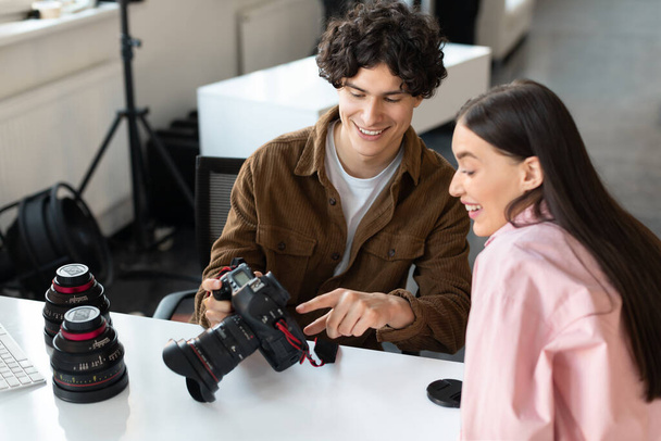 Male photographer and woman model discussing photos with camera, choosing images from photoshoot. Young cameraman talking to lady to choose the best picture, sitting in creative studio - Foto, afbeelding