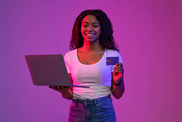 Онлайн платежі. Посмішка африканської американки з кредитною карткою і ноутбуком в руках стоять в неоновому освітленні над пурпуровим фоном, щаслива чорна жінка робить інтернет-магазин або оплачує рахунки - Фото, зображення