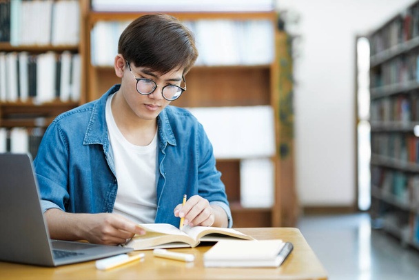 Young male university college student wearing eyeglasses and in casual cloths studying, reading, and writing down notes using laptop on desk at library for school project or exam preparation. E-Learning and Education concept. - Фото, изображение