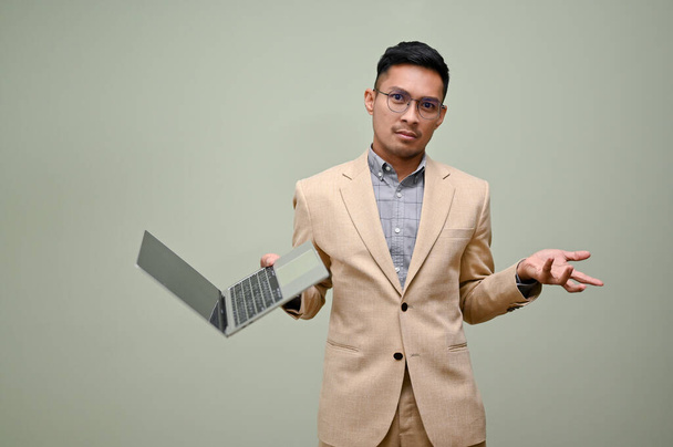 Zdezorientowany i wątpliwy milenijny azjatycki biznesmen w formalnym garniturze biznesowym stoi na tle zielonego studia z laptopem. - Zdjęcie, obraz