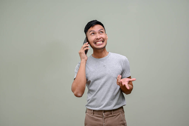 Cheerful and smiling millennial Asian man in casual clothes talking on the phone with someone, dealing or negotiating something, standing against a green studio background. - Foto, afbeelding