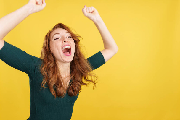Mujer pelirroja feliz celebrando mientras levanta los brazos en el estudio con fondo amarillo - Foto, Imagen