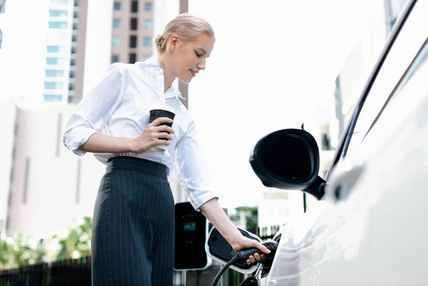 Businesswoman drinking coffee, leaning on electric vehicle recharging at public charging station with residential apartment condos building in background as progressive lifestyle by eco-friendly car. - Φωτογραφία, εικόνα