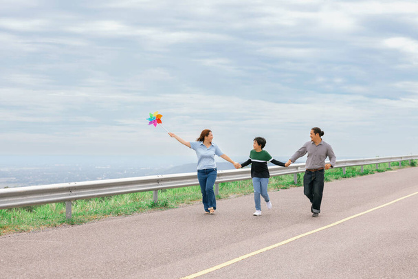 Progressive happy family vacation and carefree day concept. Young parents mother father and son run along and flying kite together road with enjoy natural scenic on scenery and clear sky background. - Photo, image