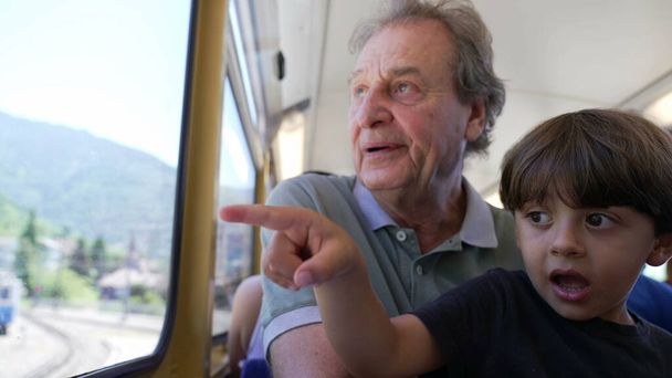 Grandfather traveling by train with grandchild seated by window looking at scenery. Senior man bonding with baby child together. Generational concept - 写真・画像