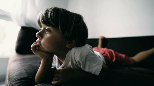 Ein kleiner Junge liegt auf dem Sofa, die Hand im Kinn, hypnotisiert von den Unterhaltungsmedien zu Hause drinnen. Lifestyle-Aufnahme eines Kindes beim Zeichentrickgucken - Foto, Bild