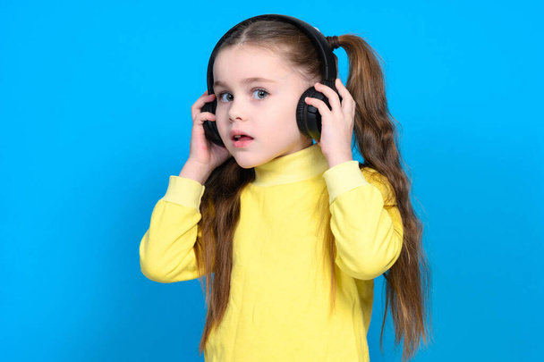 A little girl in a yellow sweater on a blue background listens to music in wireless headphones and holds them in her hands. - Fotó, kép