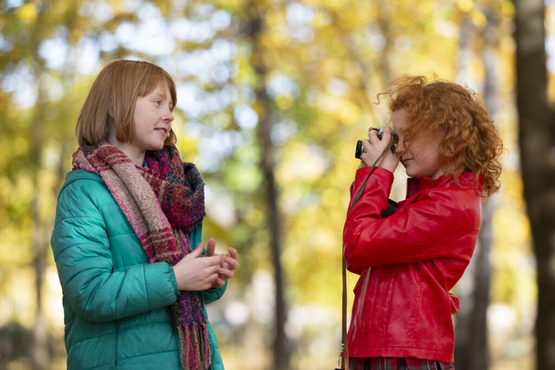 2人の赤い髪の女の子のガールフレンドは、レトロなカメラで秋の公園でお互いを写真. - 写真・画像