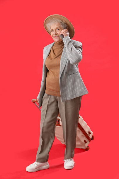 Senior woman with suitcase talking by mobile phone on red background - Fotoğraf, Görsel
