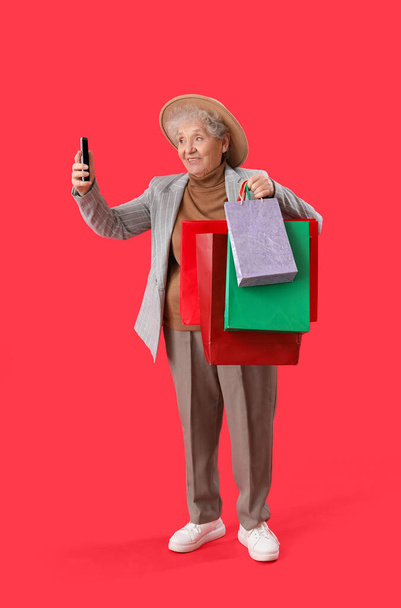 Kırmızı arka planda cep telefonu ve alışveriş torbaları olan yaşlı bir kadın. - Fotoğraf, Görsel