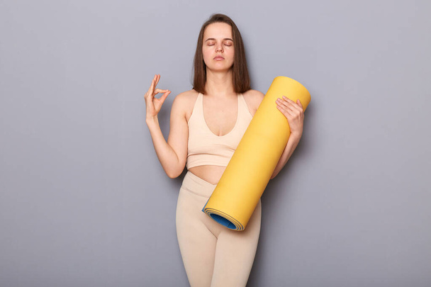 Indoor shot of calm woman with good body shape wearing beige top and leggins holding fitness mat isolated over gray background, meditating, practicing yoga with closed eyes - Fotó, kép