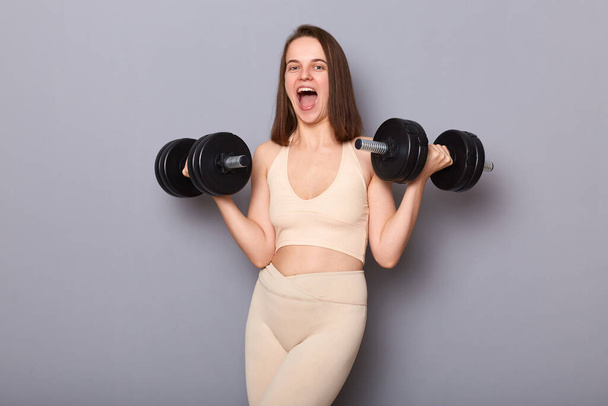Horizontal shot of overjoyed sporty woman wearing sportswear working out with weights isolated on gray background, holding two black barbels, screaming with excitement, enjoying her training - Photo, Image