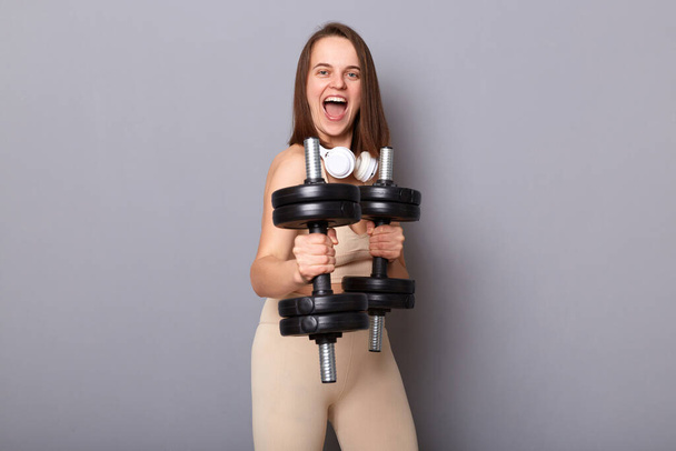 Horizontal shot of happy crazy funny woman wearing sportswear working out triceps and biceps with weights, showing dumbbells, screaming, has work out for her arms, posing isolated on gray background - Fotó, kép