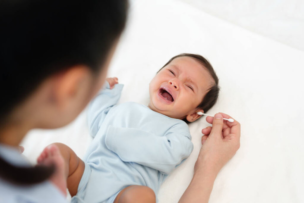 мать, использующая хлопчатобумажный бутон для очистки уха плачущего новорожденного ребенка на кровати - Фото, изображение