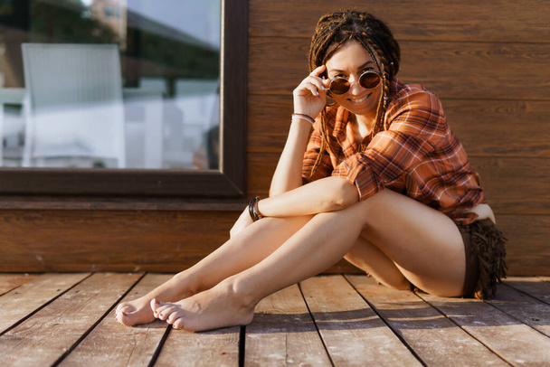 una ragazza con un'acconciatura dreadlocked posa sulla spiaggia in estate contro una parete di legno marrone rosso, luce solare brillante, vestita con una camicia a quadri e pantaloncini - Foto, immagini