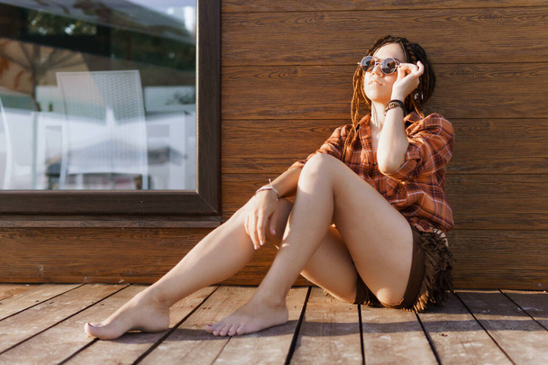 a girl with a dreadlocked hairstyle poses on the beach in summer against a red brown wooden wall, bright sunlight, dressed in a plaid shirt and shorts - Photo, Image