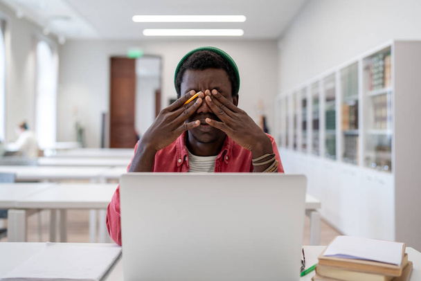 Измученный афроамериканец сидит за библиотечным столом с ноутбуком и чувствует себя нехорошо, протирая сухие раздражительные глаза, пытаясь сконцентрироваться. Уставший чернокожий фрилансер с мигренью напряжения после компьютерной работы - Фото, изображение
