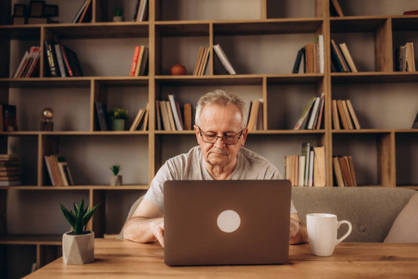 Close up mature man wearing headphones using laptop, making video call, sitting at table in kitchen, senior teacher mentor wearing glasses engaged online conference, recording webinar, teaching - Фото, изображение