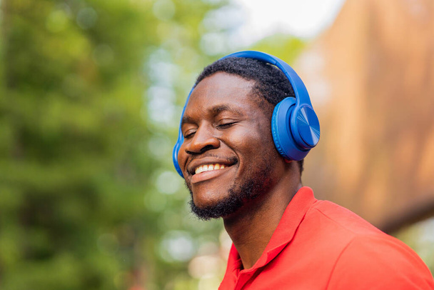 african american man in red t-shirt listening music in blue headphones and being in high spirit outdoors . - Foto, afbeelding