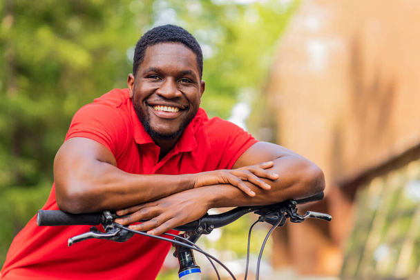 latynoski amerykański mężczyzna w luźnych ubraniach uśmiecha się opierając się na rowerze. - Zdjęcie, obraz