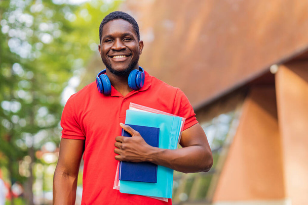 young afro american man with headphones on neck holding book and folder in hands in summer park. - Photo, Image